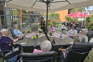 Mehrere ältere Menschen sitzen im Garten an einem rechteckigen Tisch unter einem großen Sonnenschirm und unterhalten sich