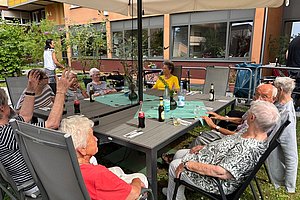 Ältere Menschen sitzen im Garten an einem rechteckigen Tisch unter einem Sonnenschirm und unterhalten sich