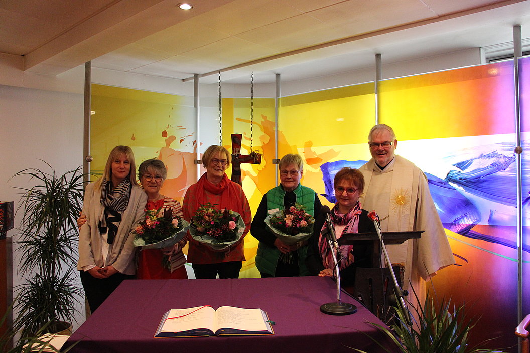 Sechs Personen stehen hinter einem Altar mit aufgeschlagenem Buch, vor einer farbigen Wand mit abstrakten Mustern.