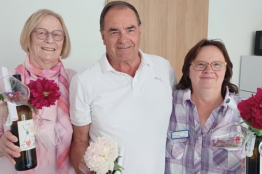 Drei Personen stehen nebeneinander, zwei Frauen halten rote Blumen und eine Flasche, der Mann in der Mitte hält eine weiße Blume