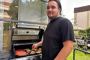 Mann in schwarzem T-Shirt steht an einem Gasgrill und wendet mit einer Grillzange Würstchen und Burgerpatties.