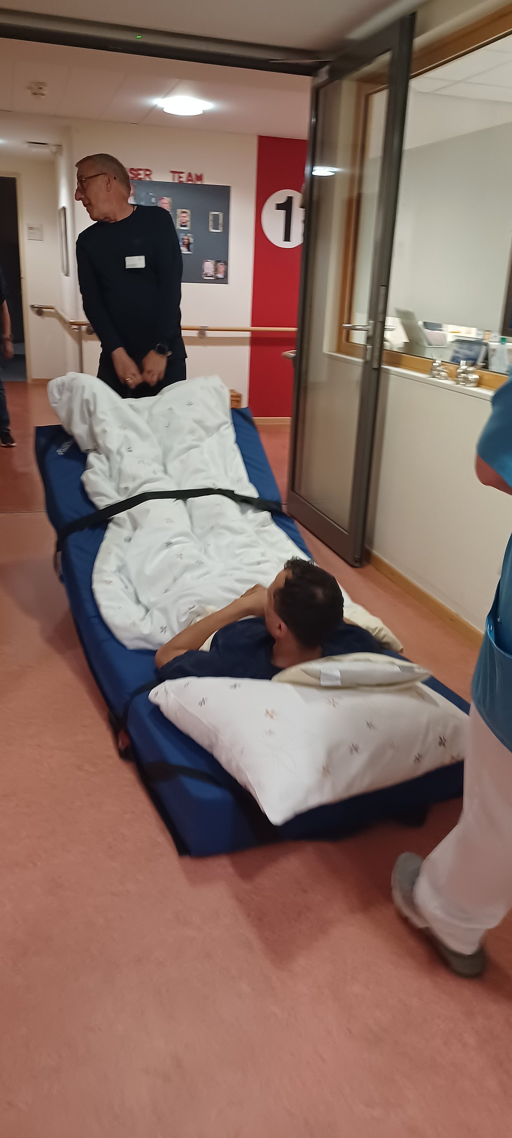 Person liegt auf einer Trage mit Decke und Kissen, umgeben von mehreren Personen in einem Flur.