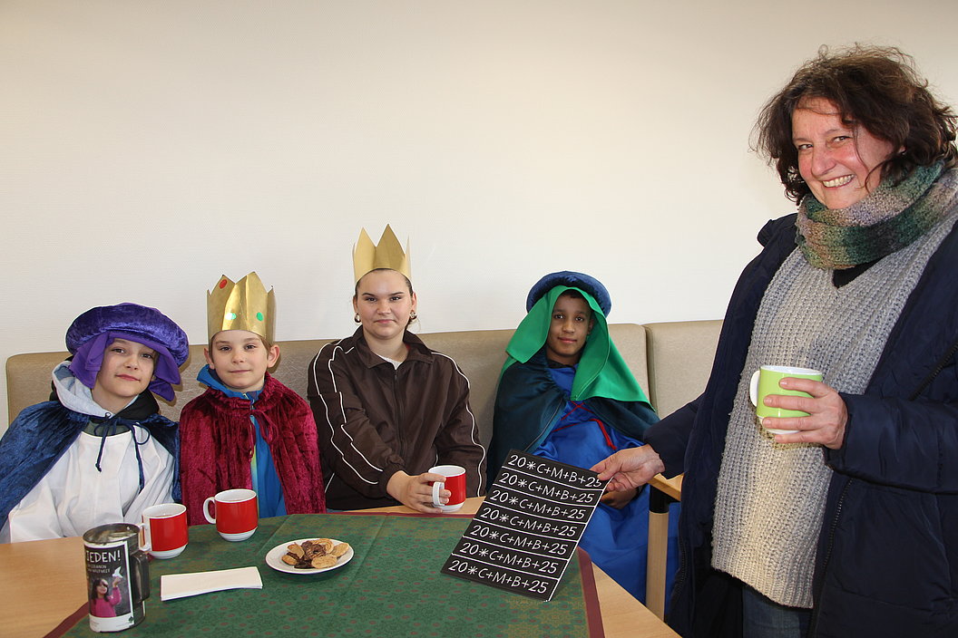 Vier Kinder in bunten Kostümen sitzen an einem Tisch mit Tassen und Keksen, eine Person steht daneben und hält eine Tafel mit mathematischen Formeln.
