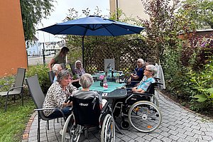 Mehrere ältere Menschen sitzen an einem Gartentisch unter einem blauen Sonnenschirm, zwei davon im Rollstuhl, umgeben von Pflanzen und Gebäuden.