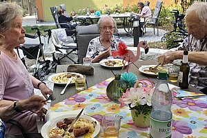 Mehrere ältere Menschen sitzen an einem mit Blumenmuster bedeckten Gartentisch und essen gemeinsam im Freien