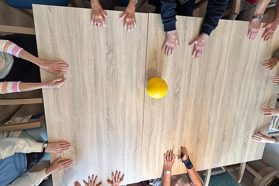 Tischrunde mit Händen von Teilnehmern und einem gelben Ball in der Mitte auf einem hellen Holztisch.