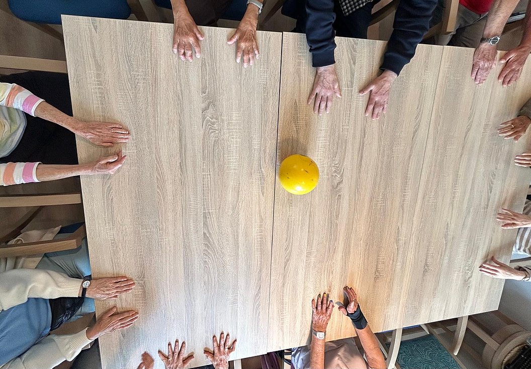 Tischrunde mit Händen von Teilnehmern und einem gelben Ball in der Mitte auf einem hellen Holztisch.