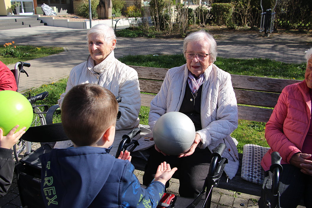 Ein Junge reicht einer älteren Frau auf einer Parkbank einen Ball, umgeben von weiteren älteren Personen.
