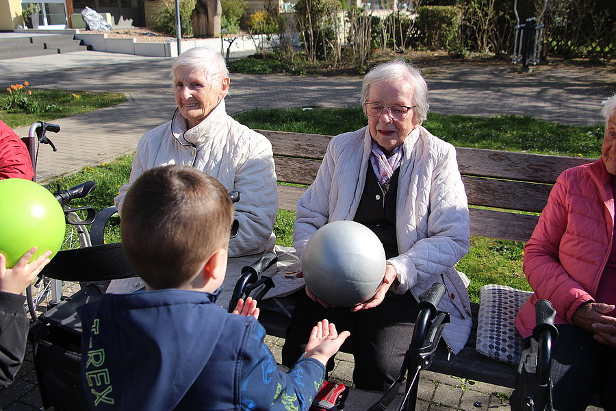 Ein Junge reicht einer älteren Frau auf einer Parkbank einen Ball, umgeben von weiteren älteren Personen.