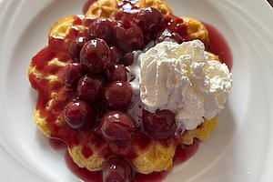 Waffel mit Kirschsoße, ganzen Kirschen und einem Klecks Schlagsahne auf weißem Teller