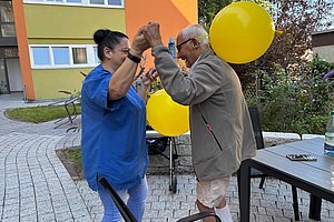 Eine Pflegerin hält die Hände eines älteren Mannes mit zwei gelben Luftballons im Hintergrund im Außenbereich eines Wohnhauses.