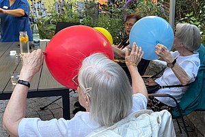 Zwei ältere Frauen sitzen im Freien an einem Tisch und halten jeweils einen aufgeblasenen Ballon vor sich, eine Frau hält einen roten, die andere einen blauen Ballon.