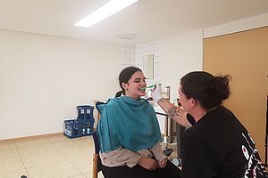 Person sitzt auf Stuhl mit Umhang, während eine andere Person ihr die Zähne putzt.