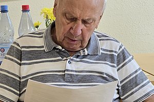 Älterer Mann mit kurz geschnittenem weißen Haar und gestreiftem Poloshirt sitzt und liest konzentriert ein Blatt Papier