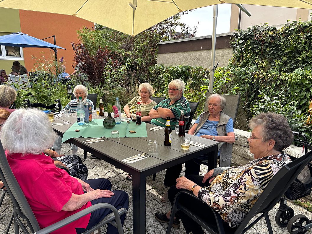 Mehrere ältere Menschen sitzen an einem rechteckigen Gartentisch unter einem Sonnenschirm im Freien und unterhalten sich