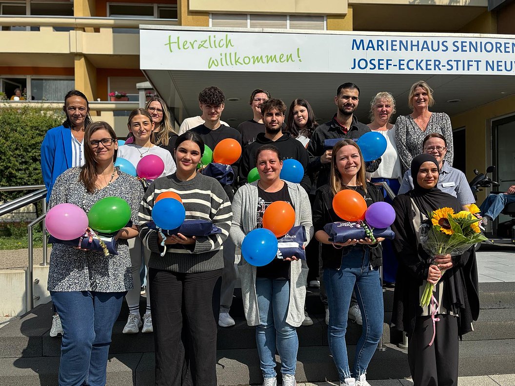 Gruppe von Menschen vor einem Gebäude mit dem Schriftzug 'Marienhaus Senioren Josef-Ecker-Stift Neu' und einem Banner mit 'Herzlich willkommen' hält bunte Luftballons und Blumen