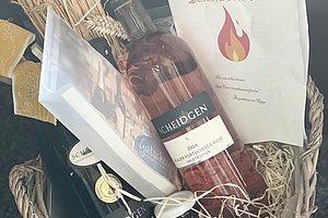 Geschenkkorb mit zwei Flaschen Wein, einem Bündel Stroh, einer Broschüre mit Flammensymbol und einem kleinen gelben Dekorationsblümchen