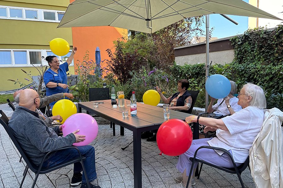 Mehrere ältere Personen sitzen an einem rechteckigen Gartentisch unter einem großen Sonnenschirm und halten bunte Luftballons