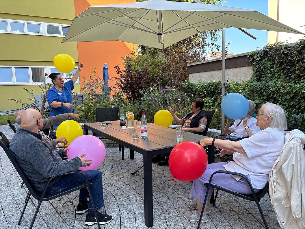 Mehrere ältere Personen sitzen an einem rechteckigen Gartentisch unter einem großen Sonnenschirm und halten bunte Luftballons