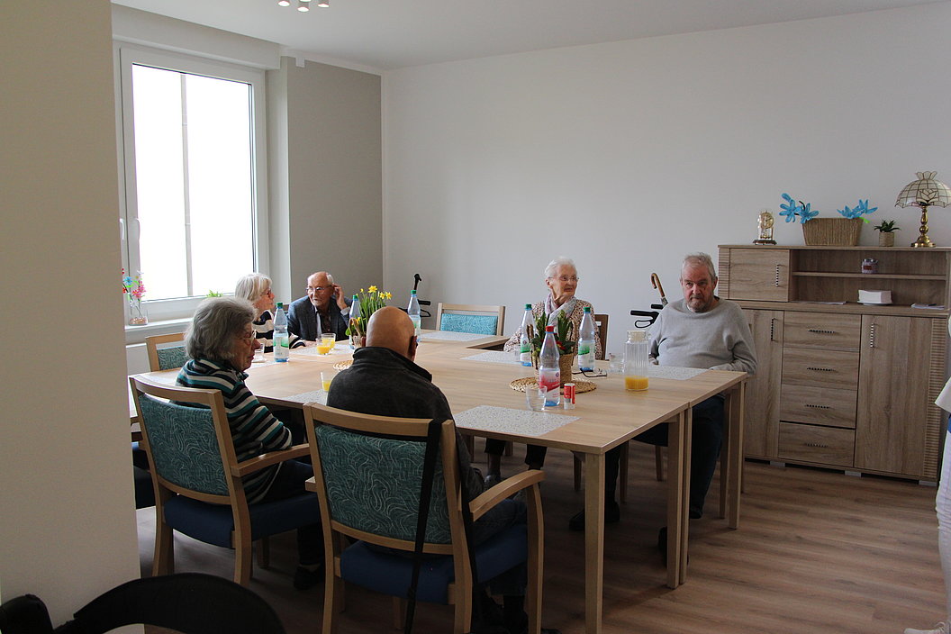 Sechs ältere Personen sitzen an einem rechteckigen Holztisch in einem hellen Raum mit Fenster und Sideboard.