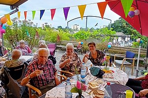 Mehrere Personen sitzen an einem langen, mit Tischdecken bedeckten Tisch unter einem Sonnenschutz mit bunten Wimpeln und einem roten Sonnenschirm im Freien.