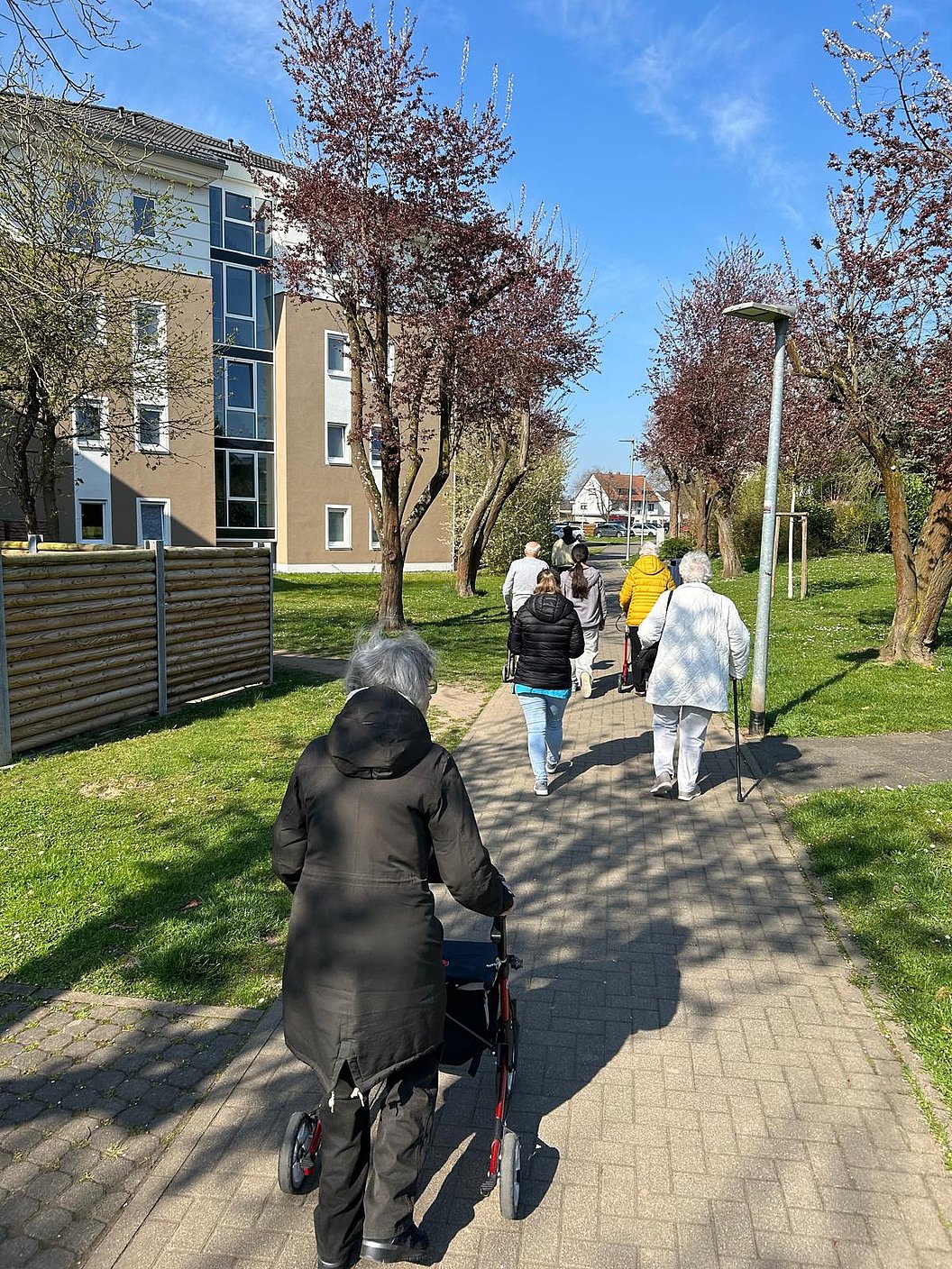 Gruppe älterer Menschen mit Jacken, die auf einem gepflasterten Weg in einem Wohngebiet spazieren gehen.