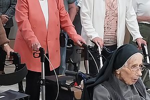 Zwei ältere Frauen mit Rollatoren stehen in einer Menschenmenge, eine weitere Person sitzt im Rollstuhl davor