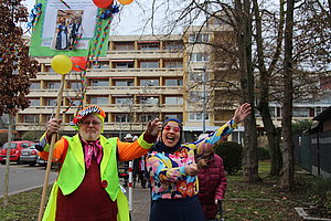 Zwei verkleidete Personen feiern fröhlich mit Luftballons und einem Banner in der Stadtstraße.
