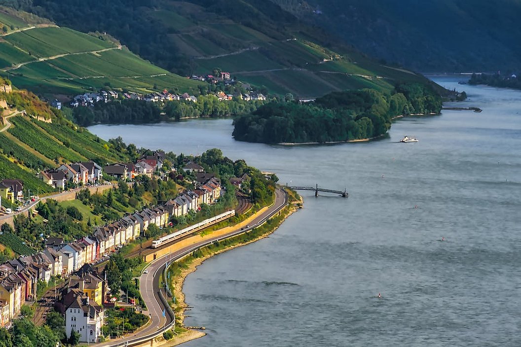 Fluss mit Uferstraße, Häusern und bewaldeten Hügeln im Hintergrund.