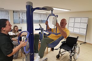 Person sitzt in einem Trainingsgerät und hält Griffe fest während eine andere Person das Gerät bedient und ein Rollstuhl daneben steht