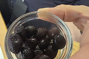 Hand hält ein Glas mit mehreren schwarzen, glänzenden Oliven