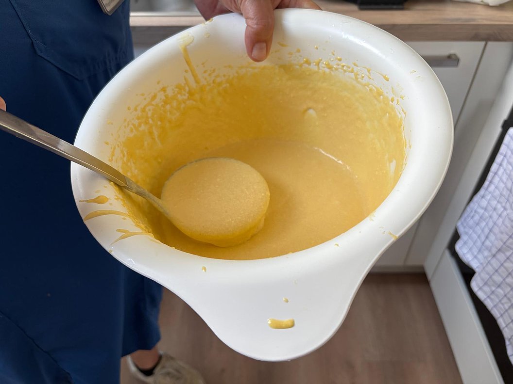 Hand hält eine weiße Schüssel mit gelbem Teig und einem Löffel darin