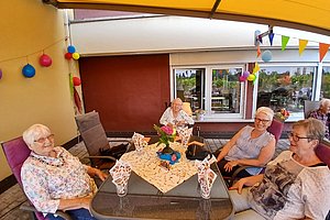 Mehrere ältere Personen sitzen an einem mit Tischdecke und Servietten gedeckten Gartentisch unter einem gelben Sonnendach auf einer Terrasse mit bunten Luftballons und Wimpeln.