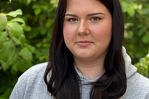Person mit langen dunklen Haaren und grauem Kapuzenpullover vor grünem Laub