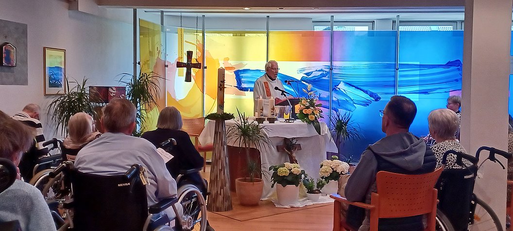 Ein Priester steht an einem mit Blumen geschmückten Altar in einem Raum mit mehreren sitzenden Menschen, einige in Rollstühlen, vor einer farbigen Glaswand mit abstraktem Muster.