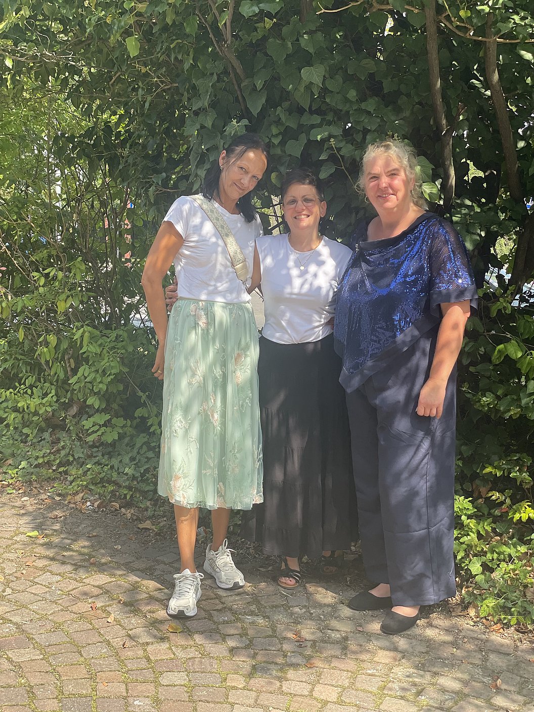 Drei Frauen stehen nebeneinander auf einem gepflasterten Weg vor dichtem Grün.