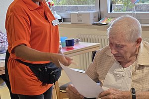 Eine Frau in orangefarbener Arbeitskleidung übergibt einem älteren Mann im Rollstuhl ein Blatt Papier in einem hellen Raum mit großen Fenstern.