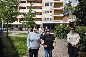 Drei Personen gehen auf einem gepflasterten Weg vor einem mehrstöckigen Wohngebäude, umgeben von Bäumen und Sträuchern.