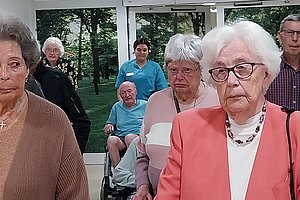 Gruppe älterer Menschen in einem hellen Raum mit großen Fenstern, einige sitzen im Rollstuhl, andere stehen oder gehen