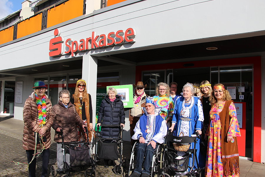 Gruppe von Menschen, einige in Rollstühlen, vor einem Sparkassen-Gebäude, einige tragen bunte, gemusterte Kleidung.