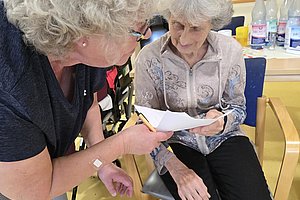 Zwei ältere Frauen sitzen an einem Tisch, eine zeigt der anderen ein Blatt Papier