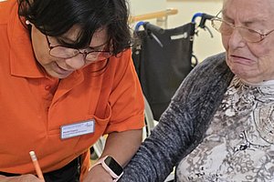 Betreuerin in orangefarbenem Poloshirt hält die Hand einer älteren Frau mit grauem Pullover und Blumenmuster und schreibt auf ein Blatt Papier