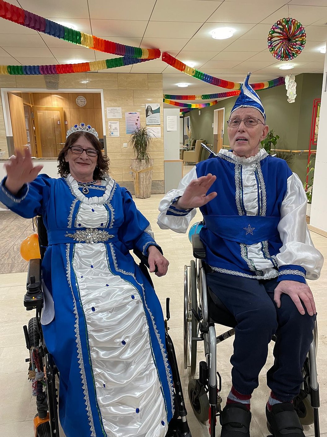 Zwei Personen in Rollstühlen, verkleidet in blauen Kostümen und Kronen, lächeln und winken.