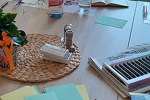 Ältere Frau sitzt an einem Tisch mit geöffnetem Buch und schreibt, umgeben von bunten Stiften, Notizzetteln und weiteren Büchern in einem hellen Raum mit Fenstern im Hintergrund