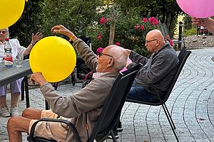 Zwei ältere Männer sitzen auf Stühlen im Freien und halten große gelbe und rosa Luftballons
