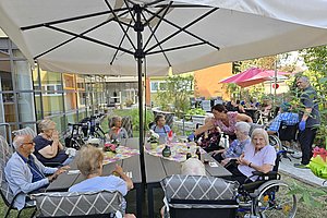 Mehrere ältere Menschen sitzen im Garten eines Pflegeheims an Tischen unter Sonnenschirmen, einige im Rollstuhl, eine Pflegerin reicht Getränke