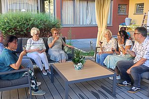 Gruppe von sechs Personen sitzt im Kreis auf Gartenmöbeln um einen niedrigen Tisch auf einer Holzterrasse
