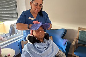 Person mit blauer Schürze und Handschuhen legt einem sitzenden Menschen ein Handtuch um den Kopf in einem Raum mit blauem Sessel und Fenster