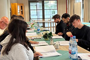 Mehrere Personen sitzen an einem langen Tisch mit grüner Tischdecke und lesen Dokumente