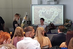 Mehrere Personen sitzen in einem Raum vor einer Leinwand, auf der ein Bild mit weißen Blumen projiziert wird.